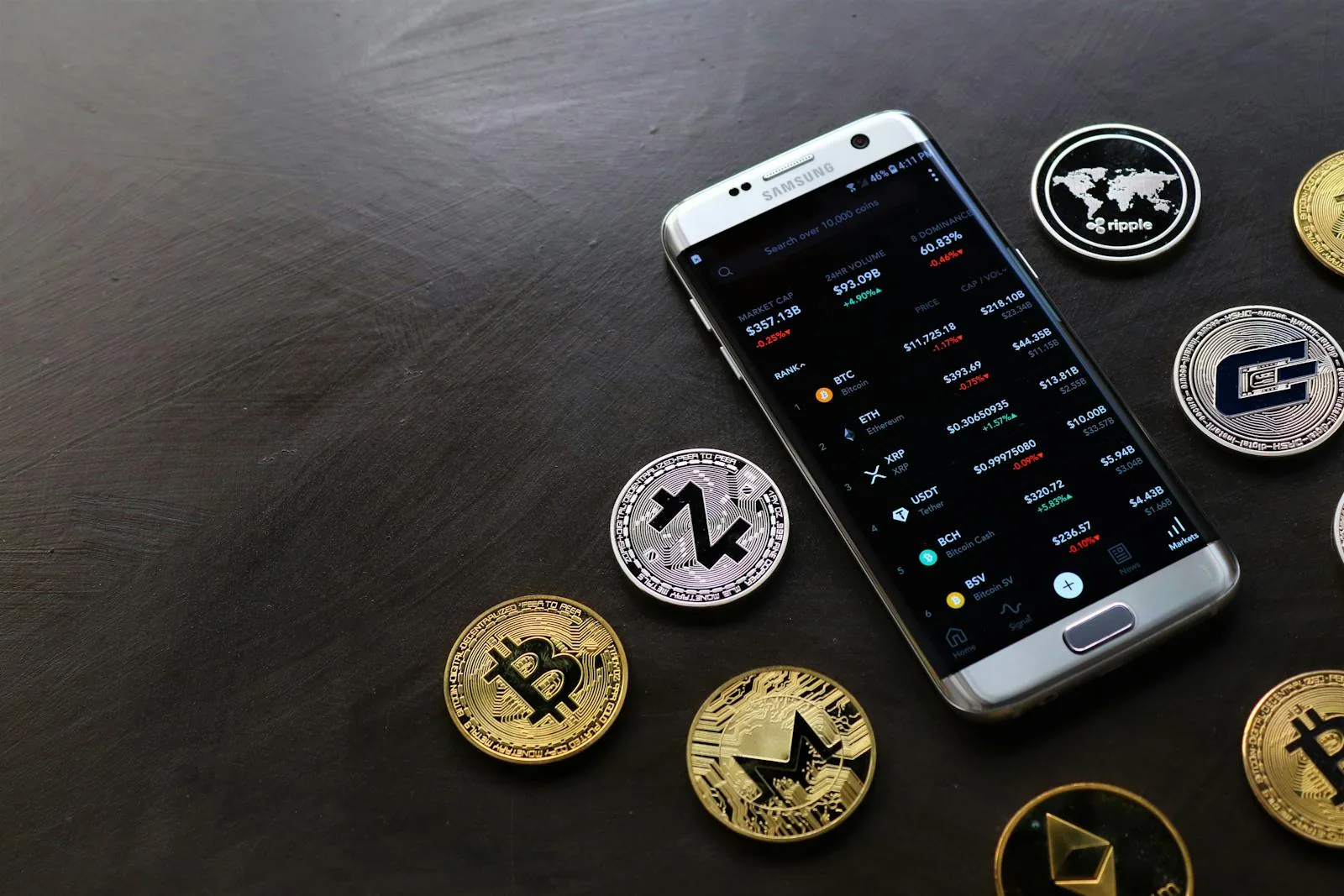 Điện thoại Samsung hiển thị ứng dụng giao dịch crypto với giá BTC ETH USDT, xung quanh là các đồng coin vật lý